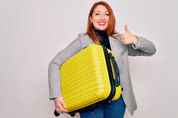 Young beautiful redhead woman holding suitcase prepared to business travel happy with big smile doing ok sign, thumb up with fingers, excellent sign