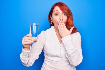 Young beautiful redhead woman drinking glass of water over isolated blue background covering mouth...