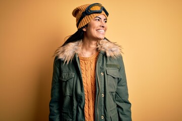 Young beautiful hispanic woman wearing ski glasses and coat for winter weather looking away to side with smile on face, natural expression. Laughing confident.