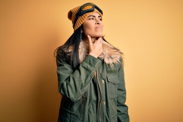 Young beautiful hispanic woman wearing ski glasses and coat for winter weather Touching painful neck, sore throat for flu, clod and infection