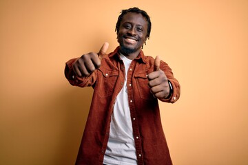 Obraz premium Young handsome african american man wearing casual jacket standing over yellow background approving doing positive gesture with hand, thumbs up smiling and happy for success. Winner gesture.