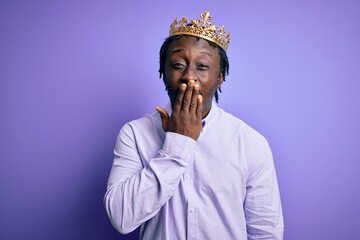 Young african american man wearing golden crown of king over isolated purple background bored yawning tired covering mouth with hand. Restless and sleepiness.