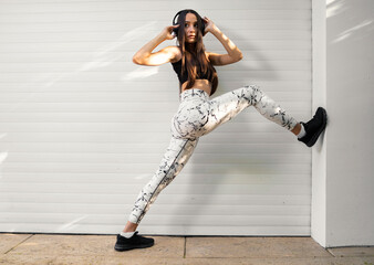 Attractive long legged athlete girl posing with headphones , stretching legs of the wall 