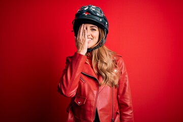 Young beautiful brunette motrocyclist woman wearing moto helmet over red background covering one eye with hand, confident smile on face and surprise emotion.
