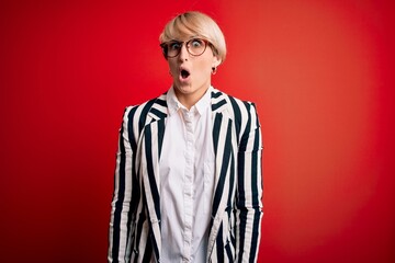 Blonde business woman with short hair wearing glasses and striped jacket over red background afraid and shocked with surprise expression, fear and excited face.