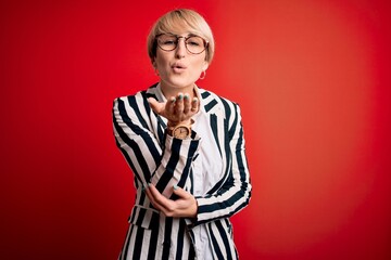 Blonde business woman with short hair wearing glasses and striped jacket over red background looking at the camera blowing a kiss with hand on air being lovely and sexy. Love expression.