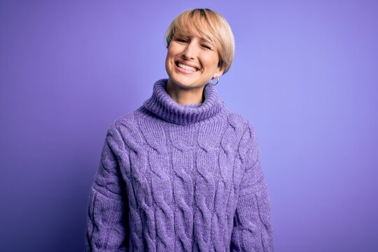 Young blonde woman with short hair wearing winter turtleneck sweater over purple background winking looking at the camera with sexy expression, cheerful and happy face.