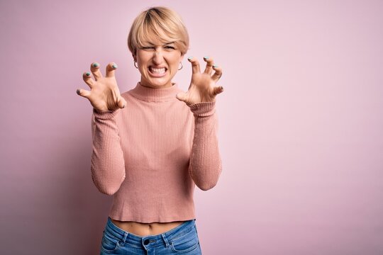 Young blonde woman with short hair wearing casual turtleneck sweater over pink background smiling funny doing claw gesture as cat, aggressive and sexy expression