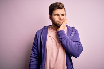 Young blond man with beard and blue eyes wearing purple sweatshirt over pink background touching mouth with hand with painful expression because of toothache or dental illness on teeth. Dentist