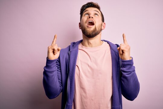 Young blond man with beard and blue eyes wearing purple sweatshirt over pink background amazed and surprised looking up and pointing with fingers and raised arms.