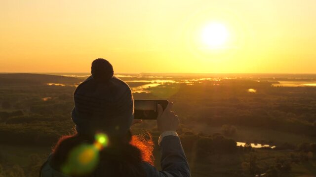 young female traveler takes photos and videos on her smartphone at dawn, from mountain in rays of beautiful sun. free girl tourist enjoys beautiful views of nature from a high hill. concept of travel