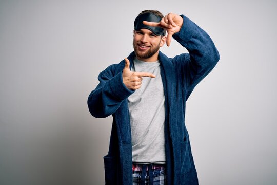 Young blond man with beard and blue eyes wearing sleep mask and pajama smiling making frame with hands and fingers with happy face. Creativity and photography concept.