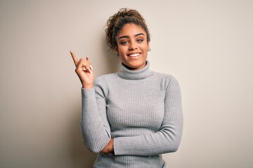 Beautiful african american girl wearing turtleneck sweater standing over white background with a big smile on face, pointing with hand and finger to the side looking at the camera.