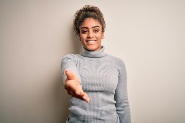 Beautiful african american girl wearing turtleneck sweater standing over white background smiling friendly offering handshake as greeting and welcoming. Successful business.