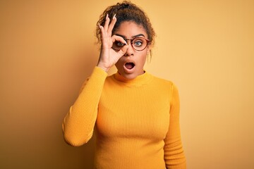 Young beautiful african american girl wearing sweater and glasses over yellow background doing ok gesture shocked with surprised face, eye looking through fingers. Unbelieving expression.