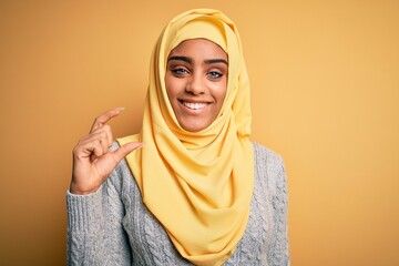 Young beautiful african american girl wearing muslim hijab over isolated yellow background smiling and confident gesturing with hand doing small size sign with fingers looking and the camera. Measure