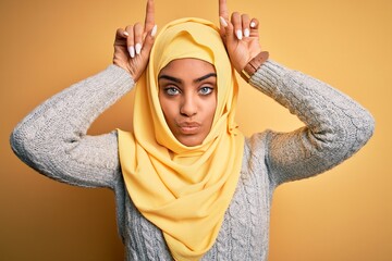 Young beautiful african american girl wearing muslim hijab over isolated yellow background doing funny gesture with finger over head as bull horns