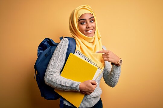 Young african american student girl wearing muslim hijab and backpack holding notebook very happy pointing with hand and finger
