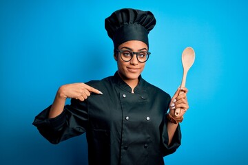 Naklejka premium Young african american chef girl wearing cooker uniform and hat holding wooden spoon tool with surprise face pointing finger to himself