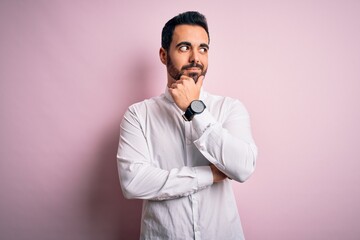 Young handsome man with beard wearing casual shirt standing over pink background with hand on chin...