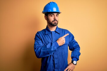 Mechanic man with beard wearing blue uniform and safety helmet over yellow background Pointing with...