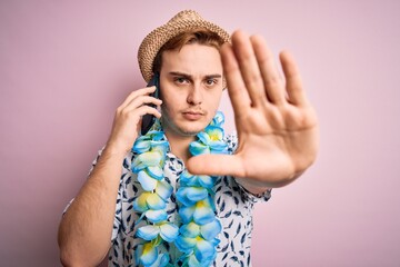 Young man on vacation wearing hat and hawaiian lei having conversation talking on smartphone with...