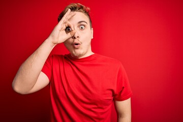 Young handsome redhead man wearing casual t-shirt over isolated red background doing ok gesture shocked with surprised face, eye looking through fingers. Unbelieving expression.