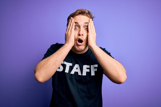 Young handsome redhead worker man wearing staff t-shirt uniform over purple background afraid and shocked, surprise and amazed expression with hands on face