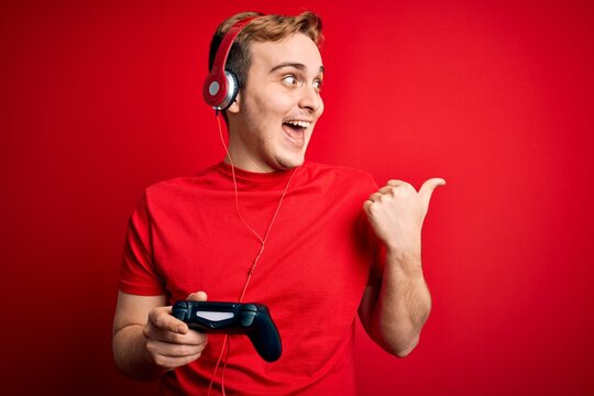 Young handsome redhead gamer man playing video game using headphones and joystick pointing thumb up to the side smiling happy with open mouth