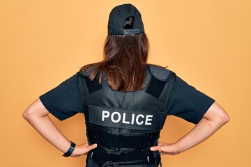 Young beautiful brunette policewoman wearing police uniform bulletproof and cap standing backwards looking away with arms on body