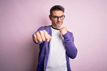Fototapeta premium Young handsome man wearing purple sweatshirt and glasses standing over pink background Punching fist to fight, aggressive and angry attack, threat and violence