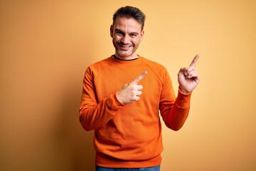 Young handsome man wearing orange casual sweater standing over isolated yellow background smiling and looking at the camera pointing with two hands and fingers to the side.