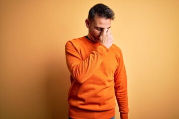 Young handsome man wearing orange casual sweater standing over isolated yellow background tired rubbing nose and eyes feeling fatigue and headache. Stress and frustration concept.