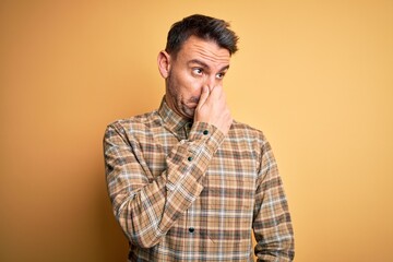 Fototapeta premium Young handsome man wearing casual shirt standing over isolated yellow background smelling something stinky and disgusting, intolerable smell, holding breath with fingers on nose. Bad smell