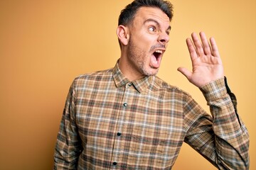 Young handsome man wearing casual shirt standing over isolated yellow background shouting and screaming loud to side with hand on mouth. Communication concept.