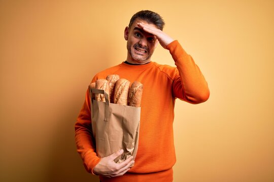 Young handsome man holding paper bag with bread over isolated yellow background stressed with hand on head, shocked with shame and surprise face, angry and frustrated. Fear and upset for mistake.