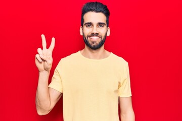 Young handsome man with beard wearing casual t-shirt smiling with happy face winking at the camera doing victory sign. number two.