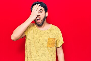 Young hispanic man wearing casual clothes peeking in shock covering face and eyes with hand,...
