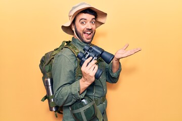 Fototapeta premium Young hispanic man wearing explorer clothes holding binoculars celebrating achievement with happy smile and winner expression with raised hand