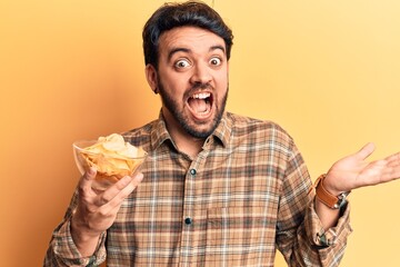 Young hispanic man holding potato chip celebrating achievement with happy smile and winner expression with raised hand