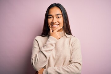 Young beautiful asian woman wearing casual turtleneck sweater over pink background looking confident at the camera smiling with crossed arms and hand raised on chin. Thinking positive.