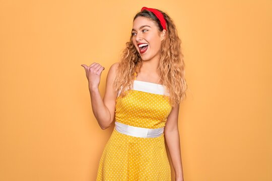Beautiful blonde pin-up woman with blue eyes wearing diadem standing over yellow background smiling with happy face looking and pointing to the side with thumb up.