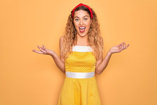 Beautiful blonde pin-up woman with blue eyes wearing diadem standing over yellow background celebrating crazy and amazed for success with arms raised and open eyes screaming excited. Winner concept