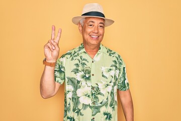 Middle age senior grey-haired man wearing summer hat and floral shirt on beach vacation showing and pointing up with fingers number two while smiling confident and happy.