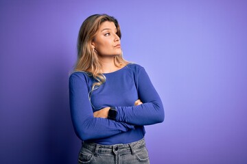 Young beautiful blonde woman wearing casual t-shirt over isolated purple background looking to the side with arms crossed convinced and confident