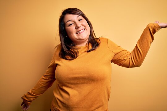 Beautiful brunette plus size woman wearing casual sweater over isolated yellow background clueless and confused expression with arms and hands raised. Doubt concept.