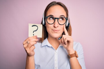 Beautiful call center agent woman working using headset holding reminder with question mark serious...