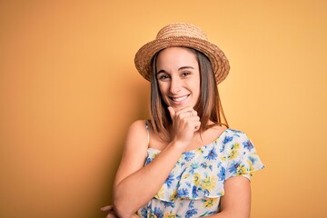 Young beautiful woman wearing casual t-shirt and summer hat over isolated yellow background looking confident at the camera smiling with crossed arms and hand raised on chin. Thinking positive.