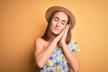 Young beautiful woman wearing casual t-shirt and summer hat over isolated yellow background sleeping tired dreaming and posing with hands together while smiling with closed eyes.