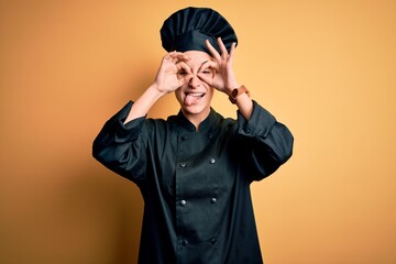 Young beautiful chef woman wearing cooker uniform and hat standing over yellow background doing ok gesture like binoculars sticking tongue out, eyes looking through fingers. Crazy expression.